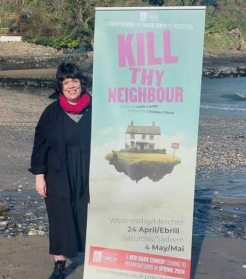 Artistic Director Chelsey Gillard with Kill Thy Neighbour play advertisement on Pembrokeshire beach.