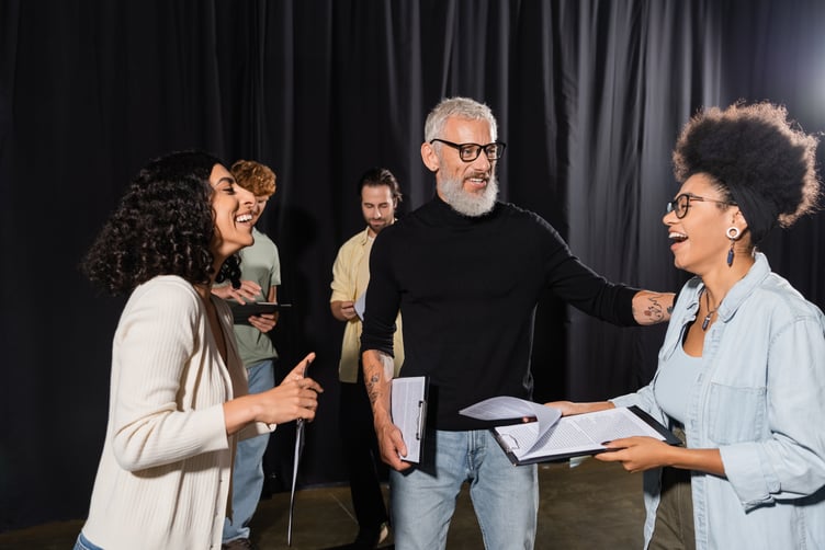 cheerful african american woman holding screenplay and laughing near bearded producer and multiethnic actors in theater,stock image