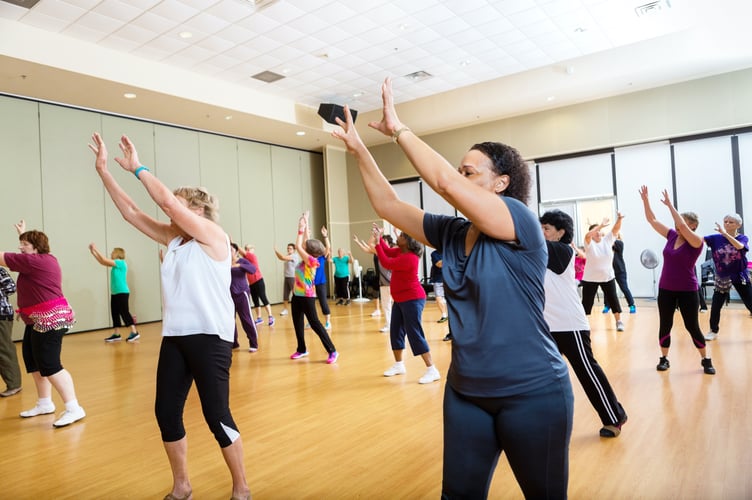 A large gorup of people are reaching up as they learn new dance moves at their class at health club. They are in the studio of the gym. The people are wearing sports clothing.